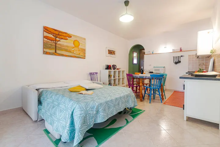Divano letto preparato come matrimoniale nella zona giorno, con cucina e tavolo sullo sfondo.